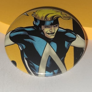 May include: A round button featuring a comic book character with blond hair wearing a blue and white costume. The character has a serious expression on their face.