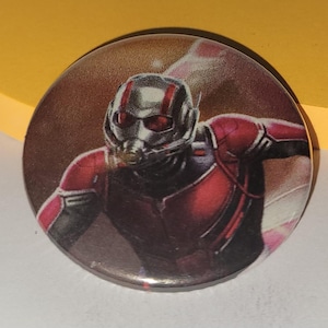 May include: Round button featuring the Ant-Man character from the Marvel comics. The button has a glossy finish and depicts Ant-Man in his red and black suit with his helmet. The background is a gradient of brown and pink.