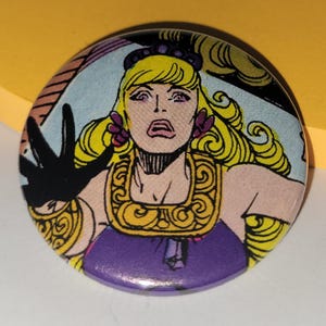 Puede incluir: Un botón redondo con una ilustración de cómic de una mujer con cabello rubio, vestida con un vestido morado y joyas doradas. La mujer tiene una expresión de sorpresa y lleva un guante negro.