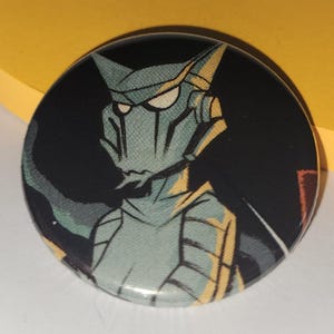 May include: Round button with a graphic of a robot-like character with a teal face and yellow accents against a black background. The character has a pointed head and angular features. The button is a collectible item.