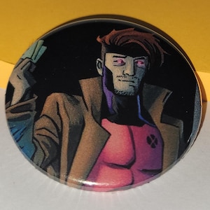 Può includere: Un bottone rotondo con un'illustrazione a fumetti di un personaggio con capelli castani, un cappotto marrone e un petto rosa. Il personaggio ha un pizzetto ed è ambientato su uno sfondo nero. Il logo degli X-Men è visibile.