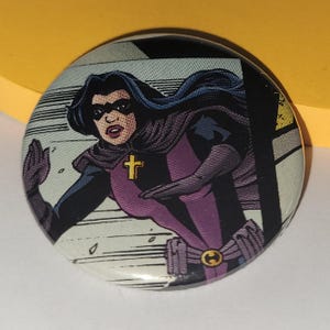Op de afbeelding: Ronde knoop met een stripboekillustratie van een vrouwelijke superheld. Ze draagt een zwart masker, cape en handschoenen, met een paars en zwart kostuum. Een gouden kruis staat op haar borst. De achtergrond heeft stripboeklijnen.