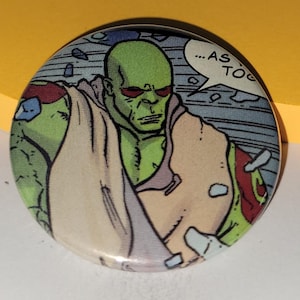 May include: Circular button with a comic book style illustration of a green-skinned character with red eyes. The character wears a tan garment and has a speech bubble with the text "...AS TO". The button has a glossy finish.