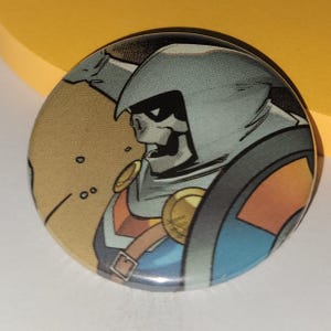 Puede incluir: Un botón redondo con un personaje de cómic con cara de esqueleto, que lleva una capa con capucha gris y una armadura azul y naranja. El personaje sostiene un escudo.