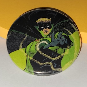 May include: Circular button with a comic book superhero illustration. The character is in a green and black costume, black cape, blonde hair, and a mask. The button has a glossy finish and a vibrant colour scheme.