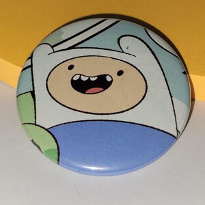 May include: A circular badge depicting a cartoon character with a white hat, a tan face, and a blue shirt. The character has a large, joyful smile. The badge has a light blue and white background.