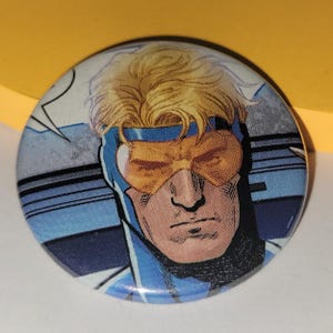 Könnte beinhalten: Runder Button mit einer Comic-Illustration eines Mannes mit blonden Haaren, gelben Brillen und einem blau-weißen Kostüm. Der Button hat ein glänzendes Finish und einen Comic-Stil.
