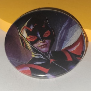 May include: A round button with a colorful image of a superhero wearing a black and red costume with a red mask. The superhero has red eyes and a serious expression.