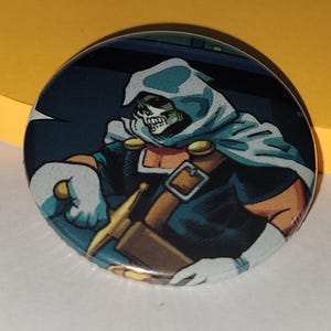 Puede incluir: Botón redondo con una ilustración de cómic de un personaje con rostro de calavera, que lleva una capa con capucha y empuña una espada. El botón tiene una combinación de colores azul, naranja y blanco, con una espada dorada.
