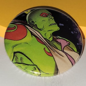 May include: A circular badge depicting a green-skinned character with red eyes and a red and orange design. The background is black and purple, with a comic book style.