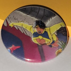 May include: Round button featuring a comic book illustration of a woman in a yellow and blue superhero costume with wings. The button has a pink and purple background with other figures in the background.