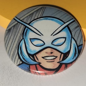 May include: Round button featuring a comic book illustration of a superhero with a blue and white mask. The button has a red and blue color scheme, with a gray background. The superhero is smiling.