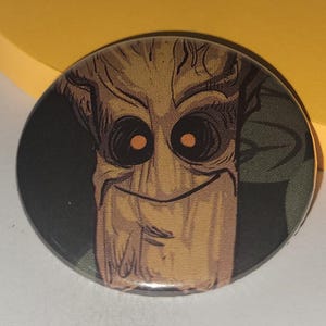 May include: A round button featuring a cartoon image of a brown tree-like character with a wide smile and orange eyes. The character is set against a dark green background.