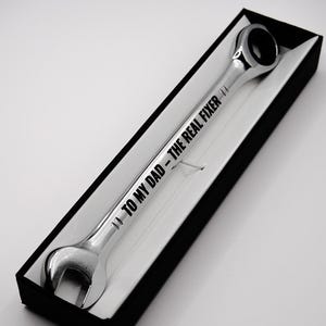 May include: A chrome wrench with the text "TO MY DAD - THE REAL FIXER" engraved on the handle. The wrench is presented in a black gift box with a white interior, ideal for a Father's Day gift or for a handyman.