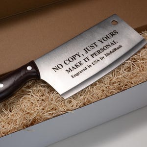 May include: A stainless steel cleaver with a dark brown handle, presented in a white box with shredded packing material. The cleaver has the text "NO COPY, JUST YOURS MAKE IT PERSONAL" engraved on the blade.