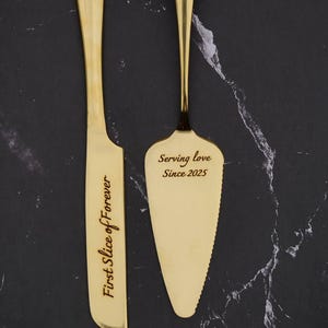 May include: A gold-colored cake knife and server set. The knife reads "First Slice of Forever" and the server reads "Serving love Since 2025". The set is displayed on a black marble-patterned surface.