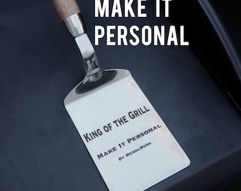May include: Stainless steel spatula with a wooden handle. The spatula has the text "KING OF THE GRILL" and "MAKE IT PERSONAL" printed on it. The spatula is on a dark surface with the words "MAKE IT PERSONAL" in large white letters.