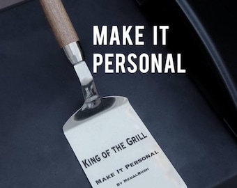 May include: Stainless steel spatula with a wooden handle. The spatula has the text "KING OF THE GRILL" and "MAKE IT PERSONAL" printed on it. The spatula is on a dark surface with the words "MAKE IT PERSONAL" in large white letters.