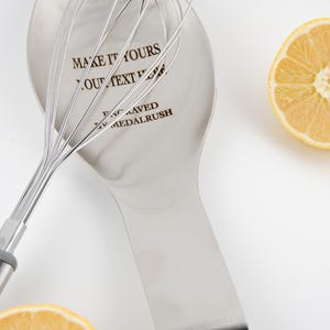May include: A stainless steel spoon rest with a whisk and a halved lemon. The spoon rest has the text "MAKE IT YOURS" and "ENGRAVED MEDALRUSH". The whisk has a gray handle. The lemon is yellow.