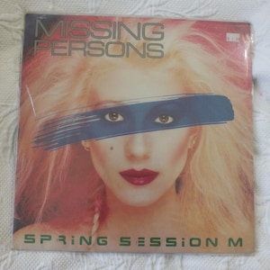 May include: A vinyl record album cover for the band Missing Persons. The cover features a woman's face with a blue stripe across her eyes. The album title is "Spring Session M". The background is a mix of pink and orange.
