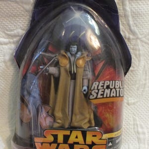 Op de afbeelding: Een Star Wars actiefiguur van een Republikeinse Senator, verpakt in een transparante plastic blister. De figuur draagt een gouden en bruine mantel. De verpakking toont het Star Wars logo en de tekst "Revenge of the Sith".
