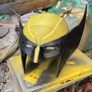 May include: A yellow and black superhero mask featuring a textured dome, pointed side fins, and white mesh eye coverings. The mask has a central yellow stripe running down the nose bridge and a distinct seam pattern across the top surface.