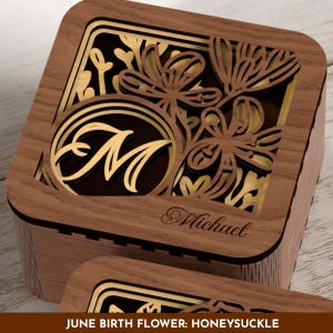 May include: Wooden box with intricate laser-cut floral design and the letter "M" in a circle. The box features the name "Michael" and the text "JUNE BIRTH FLOWER: HONEYSUCKLE". The box is made of wood with gold accents.