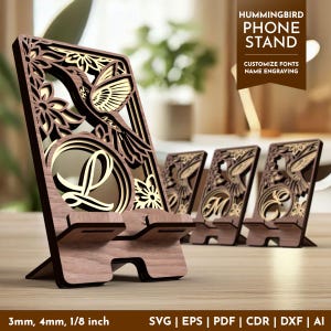 Pode incluir: Suporte de telefone de madeira com design de beija-flor cortado a laser. Apresenta um beija-flor, flores e uma letra. O texto "HUMMINGBIRD PHONE STAND" é visível. A espessura é de aproximadamente 3 mm.