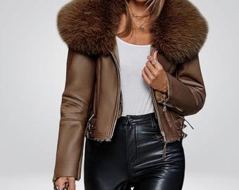 Könnte beinhalten: Eine braune Lederjacke mit einem großen, flauschigen braunen Kunstfellkragen und Innenfutter. Die Jacke verfügt über einen silberfarbenen Frontreißverschluss, lange Ärmel mit Reißverschlussbündchen und verstellbare seitliche Schnallen.