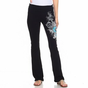 Pode incluir: Calças de ioga pretas com um design decorativo na perna inferior esquerda. O design apresenta um padrão floral com detalhes em azul e branco. A modelo está vestindo uma camisola branca.