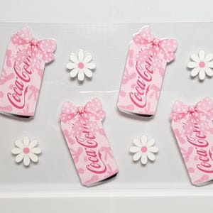 May include: Pink Coca-Cola can-shaped stickers with bows and floral patterns, featuring the Coca-Cola logo in red. White daisy-shaped stickers are also present. The stickers are arranged on a clear background.