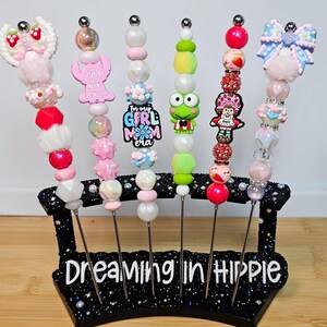 May include: A collection of beaded skewers with decorative toppers, including a frog, a girl, and a bow. The skewers are displayed in a black stand with the text "Dreaming in Hippie". The beads are in various colors, including pink, green, and red.
