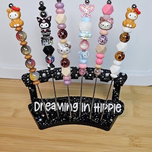 Peut inclure: Ensemble de six brochettes décoratives avec des motifs de perles et des figurines, dont Hello Kitty et un papillon. Les brochettes sont présentées dans un support noir avec le texte "Dreaming in Hippie".