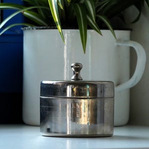 Puede incluir: Un pequeño recipiente cilíndrico de metal plateado pulido con una tapa plana y un pomo central como asa. El recipiente tiene una superficie lisa y reflectante con un diseño circular sencillo, posicionado en posición vertical.