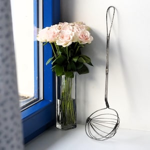 Könnte beinhalten: Ein Strauß hellrosa Rosen in einer transparenten, rechteckigen Glasvase, neben einem Vintage-Metall-Küchenschöpfer. Der Schöpfer hat einen langen Griff und einen kreisförmigen Drahtkorb-Kopf. Die Szene spielt sich in der Nähe eines blauen Fensterrahmens ab.