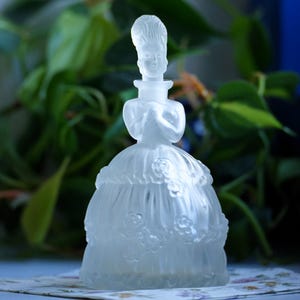 Puede incluir: Una botella de vidrio esmerilado con forma de mujer con vestido. La botella tiene un diseño detallado con detalles florales y una silueta femenina delicada.