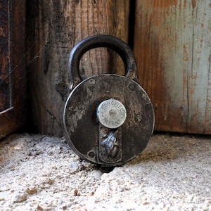 Puede incluir: Un candado redondo de hierro de estilo vintage con una pátina oscura y desgastada. El candado presenta un prominente dial central circular grabado con el texto F. SENGPIEL PATENT y un pequeño adorno de metal detallado en forma de hoja.