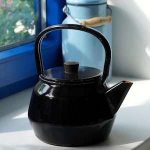May include: A black enamel tea kettle with a rounded body, a curved handle, and a matching lid featuring a small dark knob. The kettle has a short, angled spout and a smooth, glossy finish, reflecting light across its dark, solid surface.