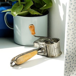 Peut inclure: Une râpe à fromage rotative vintage composée d'un tambour métallique cylindrique perforé, d'une structure en métal et de deux poignées en bois clair. Une poignée est fixée au mécanisme de manivelle, l'autre sert de poignée principale.