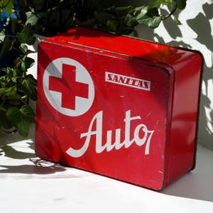 Op de afbeelding: Een vintage rode metalen EHBO-kit met een wit kruisembleem en het woord "SANITAS" boven het woord "Auto" in witte letters. De rechthoekige doos vertoont tekenen van slijtage.
