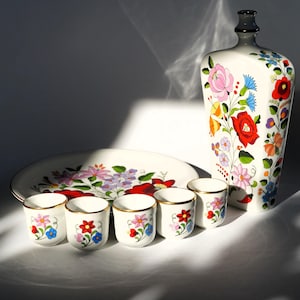 May include: A white ceramic decorative set featuring a tall bottle, a large plate, and six small cups. Each piece is adorned with colourful floral designs in red, pink, blue, and yellow. The cups have gold rims, and the set is ideal for serving drinks.