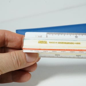 May include: A white and orange ruler with black markings and the text "Made in Czechoslovakia". The ruler is held in a hand, with a blue object in the background. The ruler has measurements in millimeters.