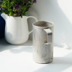 May include: A cylindrical metal mug with a matte, weathered silver finish and a simple flat handle. The mug has a straight-sided design and a plain rim. Behind the mug sits a white ceramic pitcher filled with a bouquet of purple and green flowers.