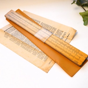 May include: A vintage wooden slide rule with clear measurement markings, on a tan leather case and aged paper documents. The slide rule is held together with a clear plastic band. The paper has text in a foreign language.