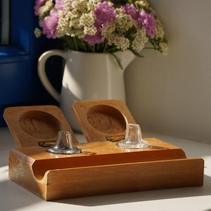 Op de afbeelding: Een rechthoekige houten bureau-organizer met twee scharnierende deksels die naar boven openen, elk met een rond verzonken compartiment. Twee kleine, doorzichtige glazen conische inzetstukken zijn geplaatst op de voorste rand van de houten basis.