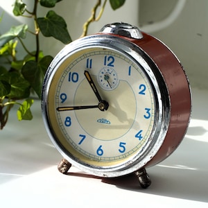 May include: A vintage alarm clock with a cream-colored face, blue numerals, and black hands. The clock has a brown and chrome casing and sits on small metal feet. The clock face has the word "PRIM" printed on it.