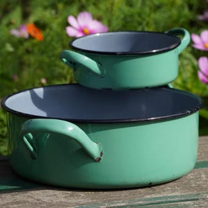 May include: Two round enamel pots in a light mint green color with black rims and side handles. The pots are stacked, with the smaller one resting on top of the larger one. Both feature a smooth, glossy finish and a light gray interior surface.