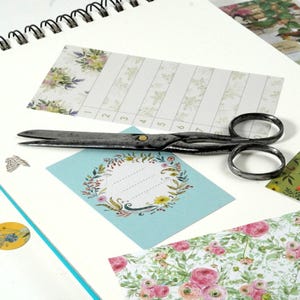 Könnte beinhalten: Eine antike Metallschere liegt auf einer weißen Oberfläche, umgeben von floral gemustertem Papier und einer hellblauen Karte mit floralem Kranzdesign. Im Hintergrund ist ein Spiralnotizbuch zu sehen. Das Bild deutet auf ein Bastel- oder Scrapbooking-Thema hin.