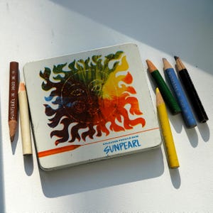 May include: A vintage Sunpearl coloured pencils set with a metal tin featuring a sun design in orange, yellow, and green. Several coloured pencils in various colours are scattered around the tin.