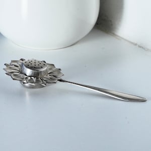 Peut inclure: Infuseur à thé argenté en forme de tournesol. L'infuseur a un centre perforé et une longue et fine poignée. Les pétales de la fleur sont détaillés et l'infuseur repose sur une surface blanche.